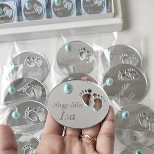 May include: Silver oval mirror with a blue gem in the centre. The mirror has a cutout of two baby footprints and the text "Hosgeldin Isa" engraved on it.
