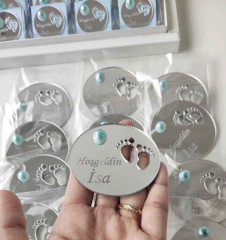 May include: Silver oval mirror with a blue gem in the centre. The mirror has a cutout of two baby footprints and the text "Hosgeldin Isa" engraved on it.