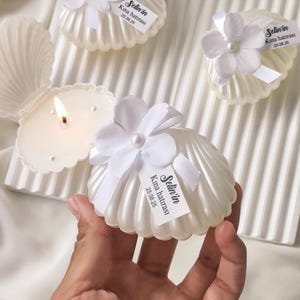 Può includere: Candele a forma di conchiglia color avorio con accenti floreali bianchi ed etichette attaccate. Le candele sono disposte su una superficie bianca strutturata, con una candela accesa. Le etichette recitano "Selin'in Kına hatırası 25.08.25".