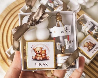 May include: A small white box with a gold ribbon and a white cloud-shaped candle. The box also contains a small wooden carousel and a chocolate bar with two bears hugging and the word "URAS" printed on it.