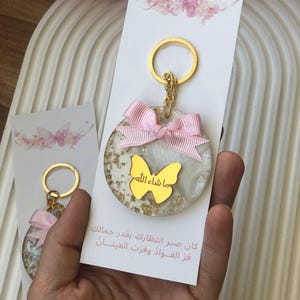 Puede incluir: Un llavero redondo con un anillo dorado y un lazo rosa. El llavero tiene un diseño de mariposa dorada con texto árabe y copos dorados. El fondo es blanco y veteado. Se adjunta una tarjeta blanca con arte de mariposas.