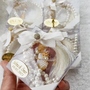 May include: White pearl prayer beads with a gold tassel and charm in a clear plastic box with a white ribbon and a gold label that says "Selma & &Ouml;mer 10.02.2021".