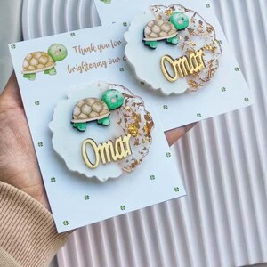 Puede incluir: Dos tarjetas con una ilustración de tortuga y el texto "Thank you for brightening our day!". Cada tarjeta tiene un adorno de resina blanca y dorada con el nombre "Omar" en letras doradas y una tortuga de madera.