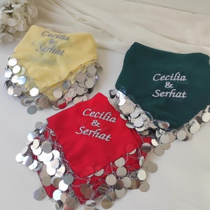 Puede incluir: Tres pañuelos cuadrados con lentejuelas plateadas alrededor de los bordes. Los pañuelos son amarillos, verdes y rojos. Cada pañuelo tiene el texto "Cecilia & Serhat" bordado en blanco.