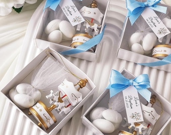 May include: Four white gift boxes tied with blue ribbons, each containing a carousel ornament, white sweets, and a tag reading "Luca's Baptism 08.23.2025". The carousel ornaments are white and gold.