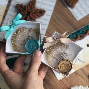 May include: Two white gift boxes with gold glitter and Arabic text. Each box has a teal or gold wax seal with a footprint design. The boxes are tied with teal and gold ribbon.