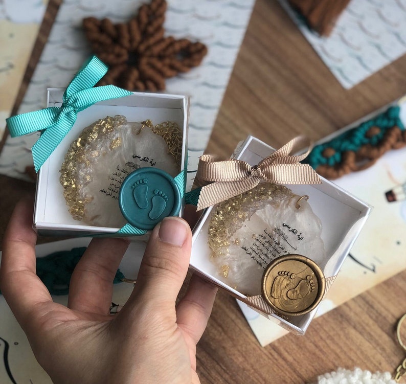 May include: Two white gift boxes with gold glitter and Arabic text. Each box has a teal or gold wax seal with a footprint design. The boxes are tied with teal and gold ribbon.