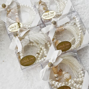 May include: Four clear plastic boxes with white ribbon bows contain white pearl prayer beads with gold accents and tassels. Each box has a gold label with the text "Selma & &Ouml;mer 10.02.2021".