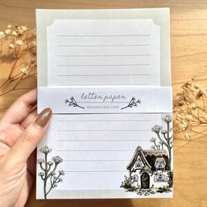 May include: A set of letter paper featuring a cottage illustration with a thatched roof and white trim. The paper has a light green border and is lined with gray lines. The text "letter paper" and "thorain.etsy.com" is printed on the front of the paper.