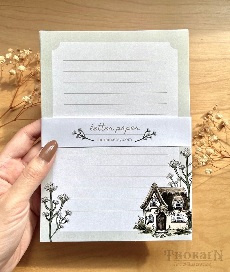 May include: A set of letter paper featuring a cottage illustration with a thatched roof and white trim. The paper has a light green border and is lined with gray lines. The text "letter paper" and "thorain.etsy.com" is printed on the front of the paper.