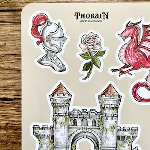 May include: A set of five stickers featuring a red dragon, a white knight's helmet with a red plume, a white rose with green leaves and stem, and a gray stone castle with a red roof and green vines. The stickers are on a beige background. The text "ThoraiN Art & Illustration" is printed in black at the top of the sheet.