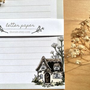 May include: A white cottage with a thatched roof and brown trim is surrounded by green foliage and white flowers. The cottage has two windows and a door. The image is on a sheet of lined paper with the text "letter paper thorain.etsy.com" at the top.