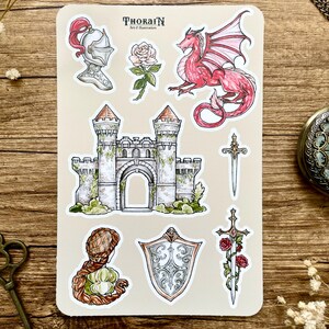 May include: A sheet of stickers featuring a castle, a dragon, a knight's helmet, a sword, a shield, and roses. The stickers are on a beige background with the text "THORAIN Art & Illustration".