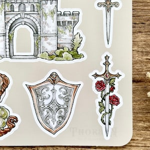 May include: A set of five stickers featuring a castle, two swords, and a shield. The castle is gray and green with a stone archway. The swords are silver with gold accents. The shield is silver with a decorative design. The stickers are on a beige background.