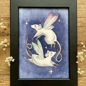 May include: A framed art print featuring two white mice with fairy wings, set against a blue background with stars. The mice are depicted in mid-flight, with one pair of wings in pink and the other in green. The artwork is enclosed in a black frame.