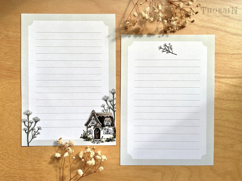 May include: Two blank lined stationery cards with a cottage illustration on one card and a floral design on the other. The cards are on a wood surface.