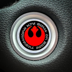 May include: A silver car steering wheel knob with a black and red Rebel Alliance symbol and the text "Rebel Alliance" in Aurebesh.