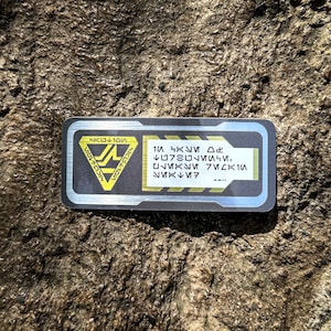 May include: A rectangular black and silver sticker with yellow accents. The sticker features a yellow triangle with a stylized logo and text in an alien language. The right side of the sticker has text in an alien language.
