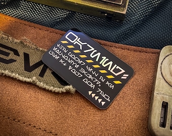 May include: A black and gold sticker with the text "OK7U104D" and a series of yellow lines. The text is in a futuristic font and includes the words "IN HU", "7N0", "DO7", "V007", "3K07", "YMN", "U00W7", "IN", and "7N0".