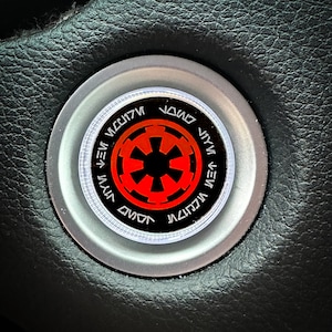 May include: A silver car steering wheel knob with a black and red Imperial symbol from Star Wars. The symbol is surrounded by a white ring with the text "JONO VEV MWCU7N JONO VEV MWCU7N" in black.