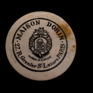 Può includere: Coperchio rotondo e bianco sporco di un antico contenitore di cosmetici Maison Dorin Paris. Il testo nero include "27 R. Grenier St. Lazare Paris" e "Marque de Fabrique". È visibile anche un emblema centrale.
