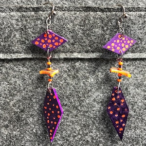Può includere: Orecchini pendenti viola e arancioni con una forma geometrica a diamante e pois arancioni. Gli orecchini sono realizzati in argilla polimerica e hanno ganci d'argento.