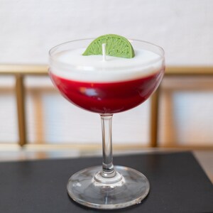 May include: A cocktail-shaped candle with a clear glass, featuring a red base, a white top, and a green lime wedge. The candle has a wick and is designed to resemble a drink.