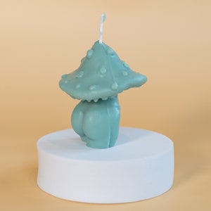 Op de afbeelding: Een teal-kleurige paddenstoelvormige kaars met een getextureerde kap en een afgeronde basis. De kaars wordt gepresenteerd op een witte, cirkelvormige sokkel tegen een lichtoranje achtergrond. De kaars heeft een lont die uit de bovenkant steekt.