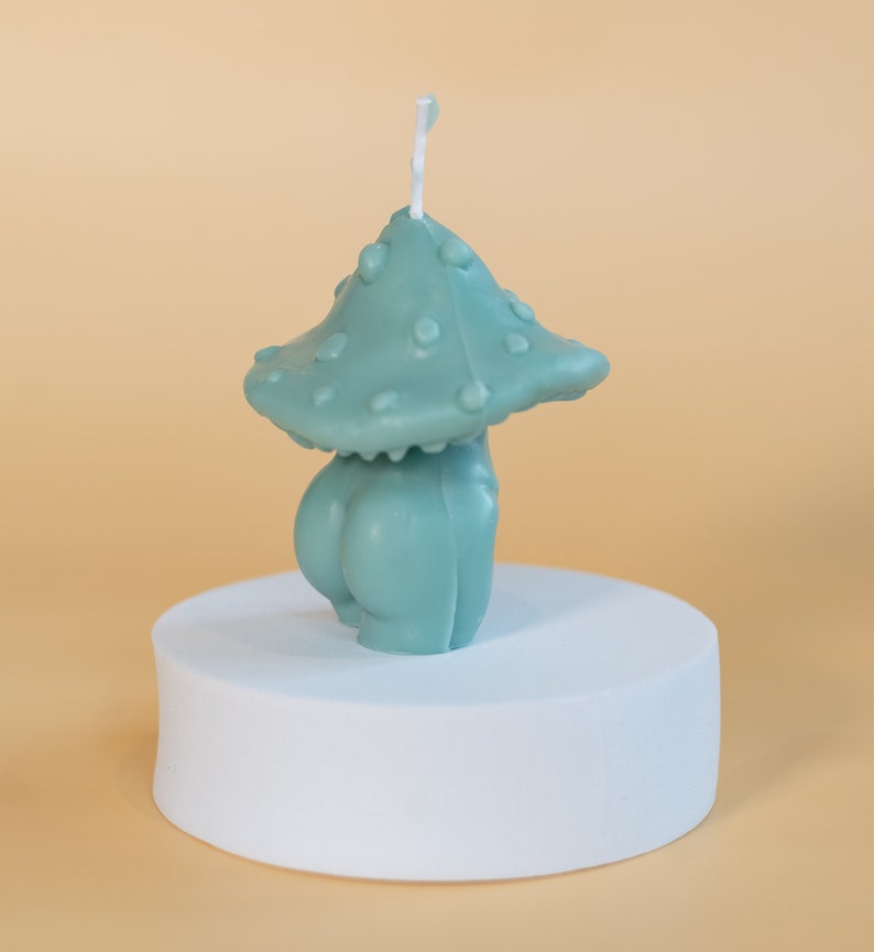 Op de afbeelding: Een teal-kleurige paddenstoelvormige kaars met een getextureerde kap en een afgeronde basis. De kaars wordt gepresenteerd op een witte, cirkelvormige sokkel tegen een lichtoranje achtergrond. De kaars heeft een lont die uit de bovenkant steekt.