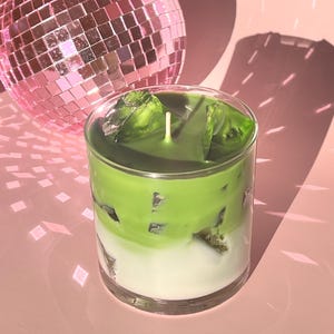 May include: A layered candle in a clear glass, featuring a white base, a vibrant green middle, and green wax chunks on top. A white wick is centered. A pink disco ball is in the background, casting light patterns on the pink surface.