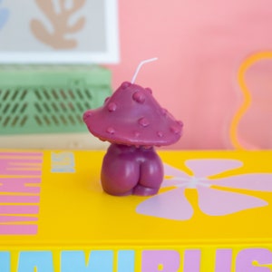 Op de afbeelding: Een grillige, paarse paddenstoelvormige kaars met een witte lont. De kaars staat op een gele ondergrond met de tekst "MIAMI BLISS" in blauw en roze. De achtergrond is zachtroze.