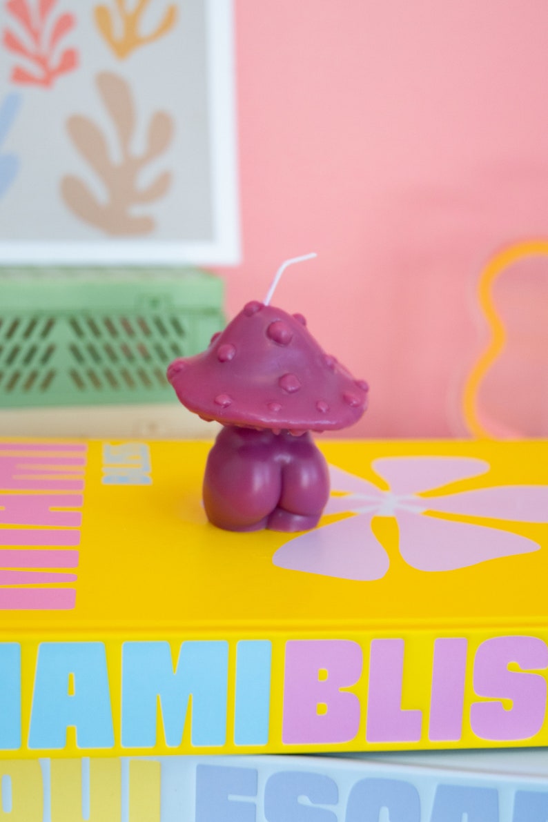 Op de afbeelding: Een grillige, paarse paddenstoelvormige kaars met een witte lont. De kaars staat op een gele ondergrond met de tekst "MIAMI BLISS" in blauw en roze. De achtergrond is zachtroze.