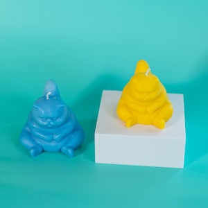 Może przedstawiać: Dwie świeczki w kształcie kota, jedna niebieska, a druga żółta, stoją na białej powierzchni. Niebieska świeczka znajduje się po lewej stronie, a żółta po prawej. Obie świeczki stoją na białej powierzchni.