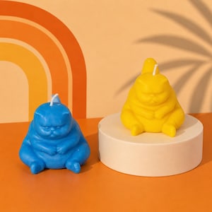 May include: Two cat-shaped candles. One is blue, the other is yellow. Both candles are sitting down with their paws crossed. The candles have a grumpy expression. The background is orange with a rainbow design.