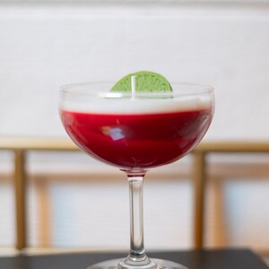 May include: A cocktail-shaped candle with a red wax base and a white top, topped with a green lime slice. The candle is in a clear stemmed glass, resembling a cocktail. The candle sits on a black surface.