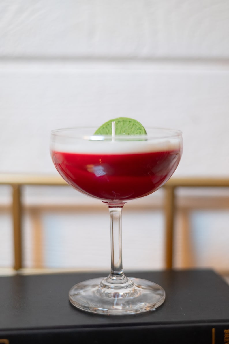 May include: A cocktail-shaped candle with a red wax base and a white top, topped with a green lime slice. The candle is in a clear stemmed glass, resembling a cocktail. The candle sits on a black surface.