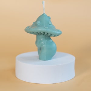 Op de afbeelding: Een teal paddenstoelvormige kaars met een getextureerde kap en een glad, afgerond lichaam. De kaars staat op een wit cilindrisch platform tegen een lichtoranje achtergrond. Een witte lont steekt uit de bovenkant van de paddenstoel.