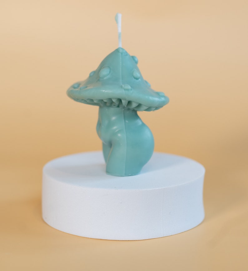 Op de afbeelding: Een teal paddenstoelvormige kaars met een getextureerde kap en een glad, afgerond lichaam. De kaars staat op een wit cilindrisch platform tegen een lichtoranje achtergrond. Een witte lont steekt uit de bovenkant van de paddenstoel.