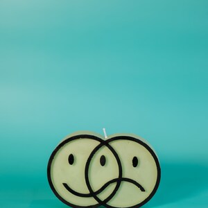 Puede incluir: Una vela verde claro con forma de dos caras sonrientes superpuestas, una feliz y otra triste, delineadas en negro. La vela tiene una mecha blanca y se encuentra sobre un fondo turquesa.