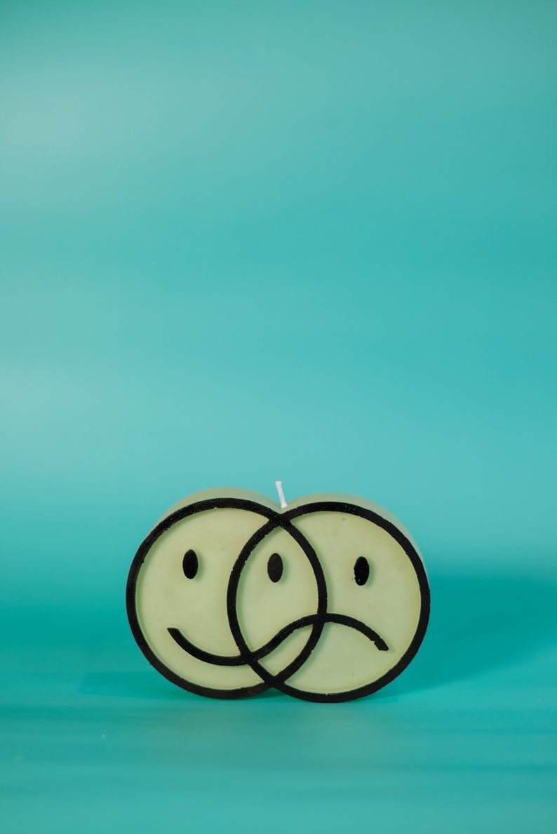 Puede incluir: Una vela verde claro con forma de dos caras sonrientes superpuestas, una feliz y otra triste, delineadas en negro. La vela tiene una mecha blanca y se encuentra sobre un fondo turquesa.