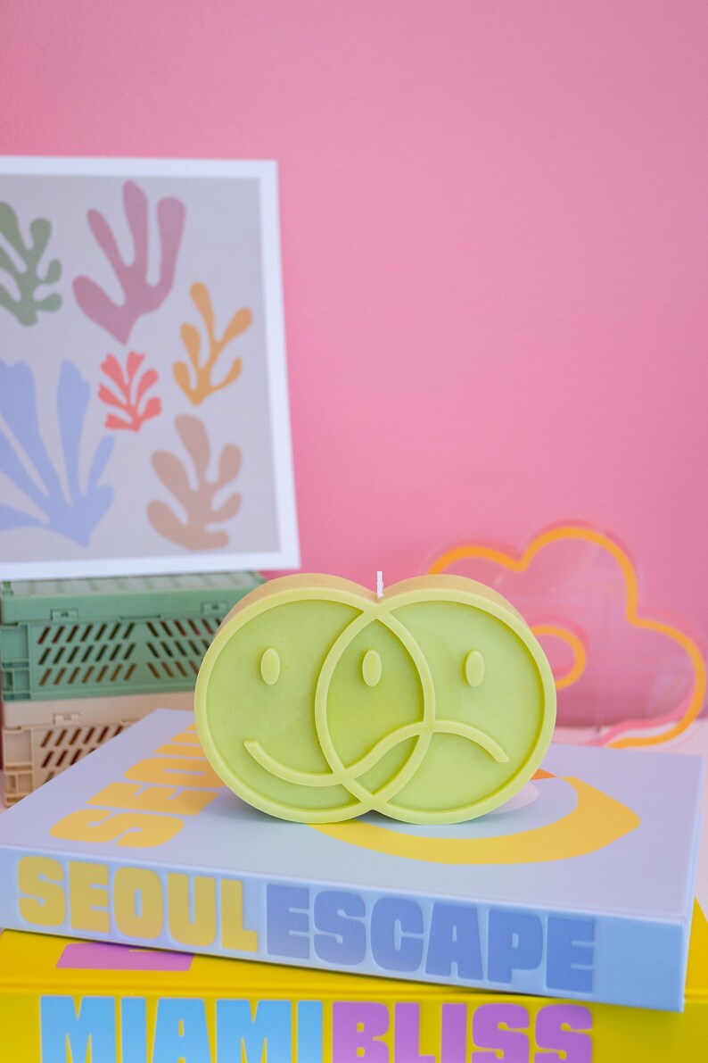 Puede incluir: Una vela verde lima con forma de dos caras sonrientes superpuestas, una feliz y otra triste. La vela est&aacute; sobre una pila de libros con los t&iacute;tulos "SEOUL ESCAPE" y "MIAMI BLISS". Una impresi&oacute;n enmarcada y una nube de ne&oacute;n est&aacute;n en el fondo.