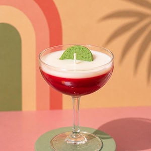 May include: A cocktail-shaped candle with a red base, white middle, and a green lime wedge on top. The candle sits in a clear coupe glass on a green pedestal. The background features a retro rainbow design.