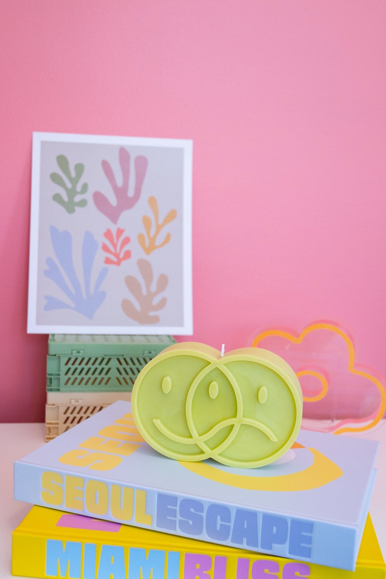 Puede incluir: Una vela verde lima con forma de dos caras sonrientes conectadas, una feliz y otra triste, descansa sobre una pila de libros. Una impresi&oacute;n enmarcada con formas abstractas coloridas y una luz de ne&oacute;n en forma de nube est&aacute;n en el fondo.