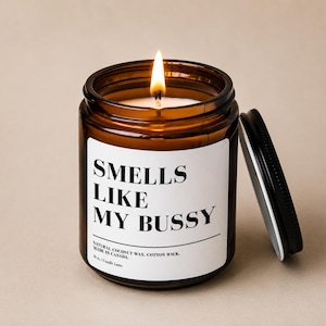 Peut inclure: Une bougie allumée dans un pot en verre ambré avec un couvercle noir. L'étiquette blanche porte le texte noir en gras SMELLS LIKE MY BUSSY, suivi de NATURAL COCONUT WAX, COTTON WICK, MADE IN CANADA, et 283g / Candle Lune.