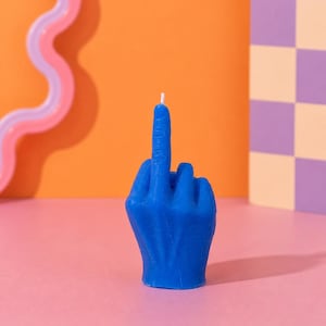 Peut inclure: Une bougie bleue en forme de main faisant un geste obscène. La bougie est posée sur une surface rose, avec un fond orange. Un objet ondulé rose et violet est à gauche, et un motif à carreaux à droite.