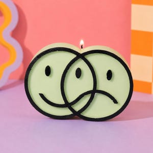 Puede incluir: Una vela verde claro con forma de dos caras sonrientes superpuestas, una feliz y otra triste, con contornos negros. La vela est&aacute; encendida, sobre un fondo rosa y naranja.