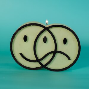 Puede incluir: Una vela &uacute;nica con forma de dos caras sonrientes superpuestas, una feliz y otra triste, con contornos negros. La vela es de color verde p&aacute;lido y tiene una mecha encendida. El fondo es de color turquesa s&oacute;lido.