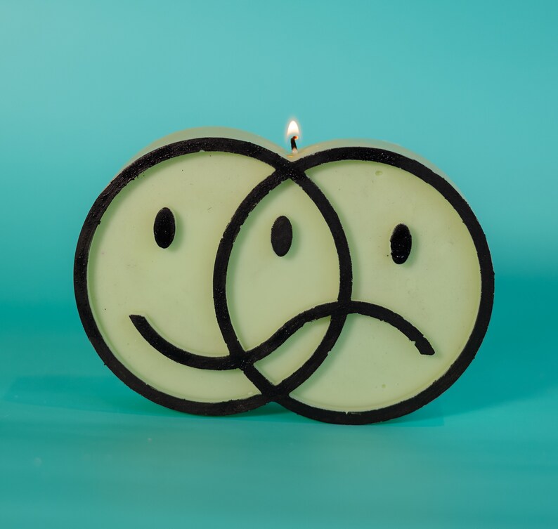 Puede incluir: Una vela &uacute;nica con forma de dos caras sonrientes superpuestas, una feliz y otra triste, con contornos negros. La vela es de color verde p&aacute;lido y tiene una mecha encendida. El fondo es de color turquesa s&oacute;lido.
