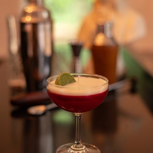 May include: A cocktail-themed candle with a layered design. The candle features a red base, a white middle, and a green lime wedge on top. Other bar tools are visible in the background, including a cocktail shaker and a mixing glass.