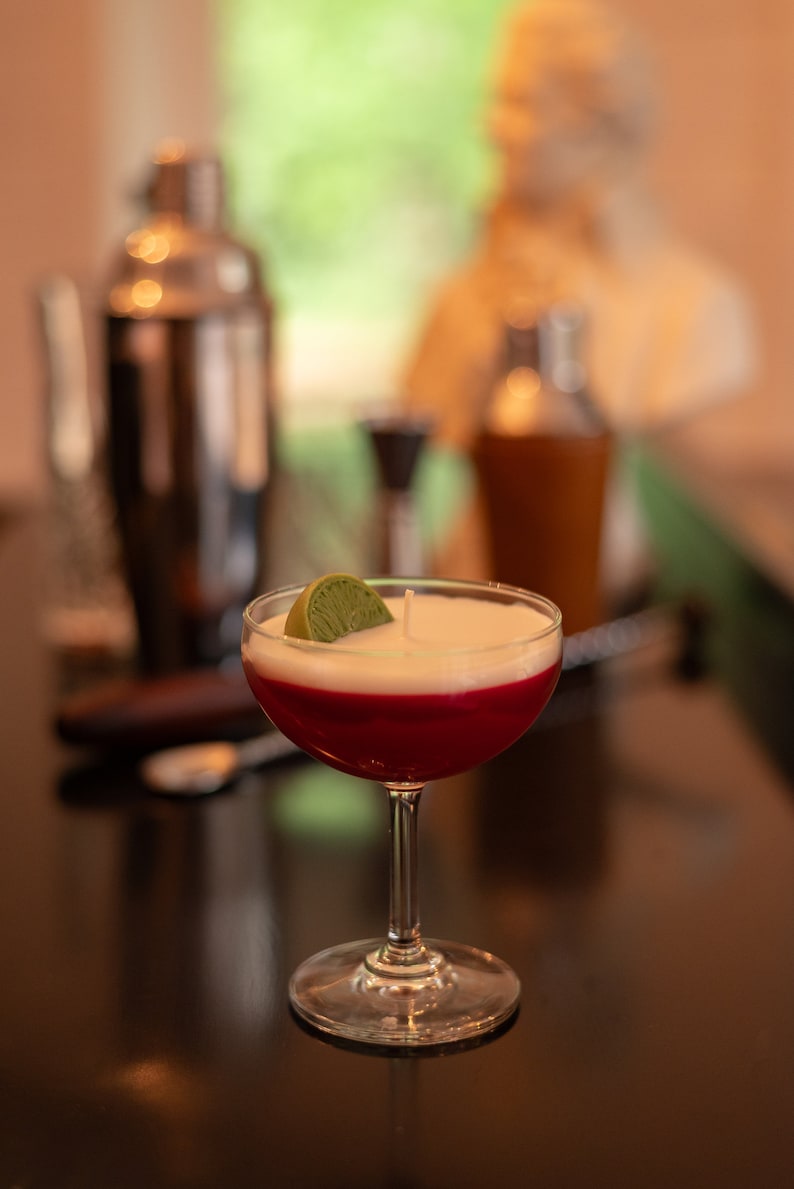 May include: A cocktail-themed candle with a layered design. The candle features a red base, a white middle, and a green lime wedge on top. Other bar tools are visible in the background, including a cocktail shaker and a mixing glass.